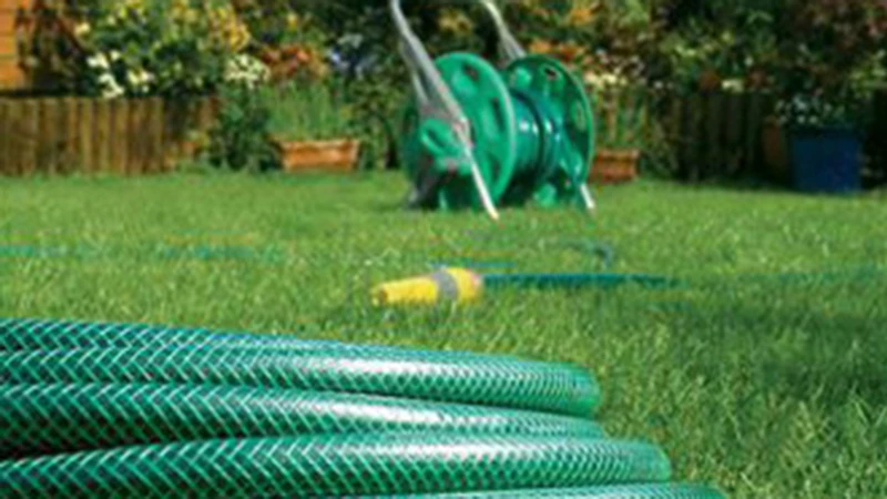 Навит зелен градински маркуч на преден план и количка с макара за маркуч в добре поддържана зелена градина.