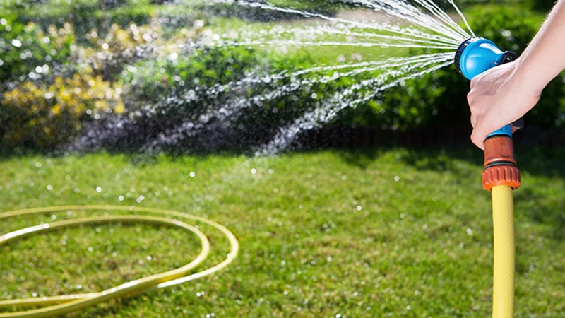 Ръка, държи градински пистолет, закрепен към жълт маркуч, който разпръсква ситна водна мъгла над зелена морава.